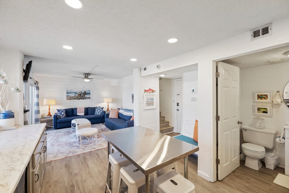 Pinnacle Port 304A – Living room with blue sofa, dining table, and bathroom; bright lighting and light wood flooring.