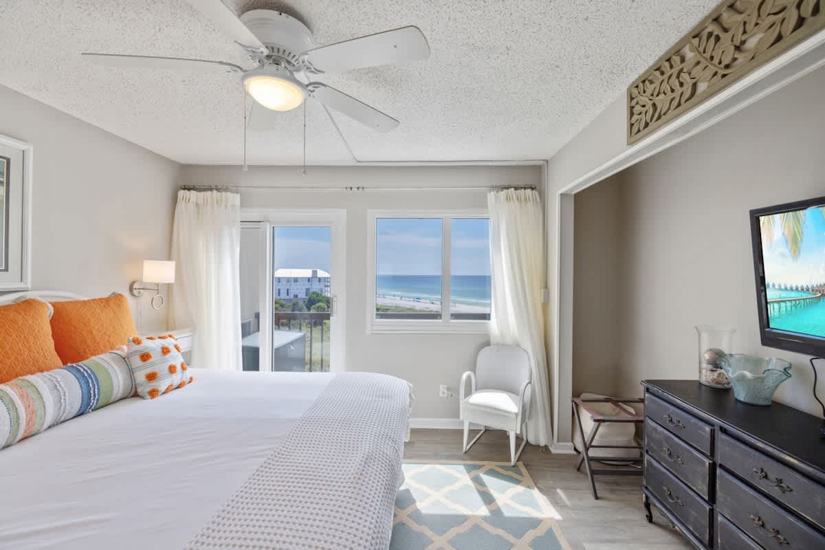 Pinnacle Port 304A – Cozy bedroom with ocean view, large windows, light curtains, and a dresser with a television.