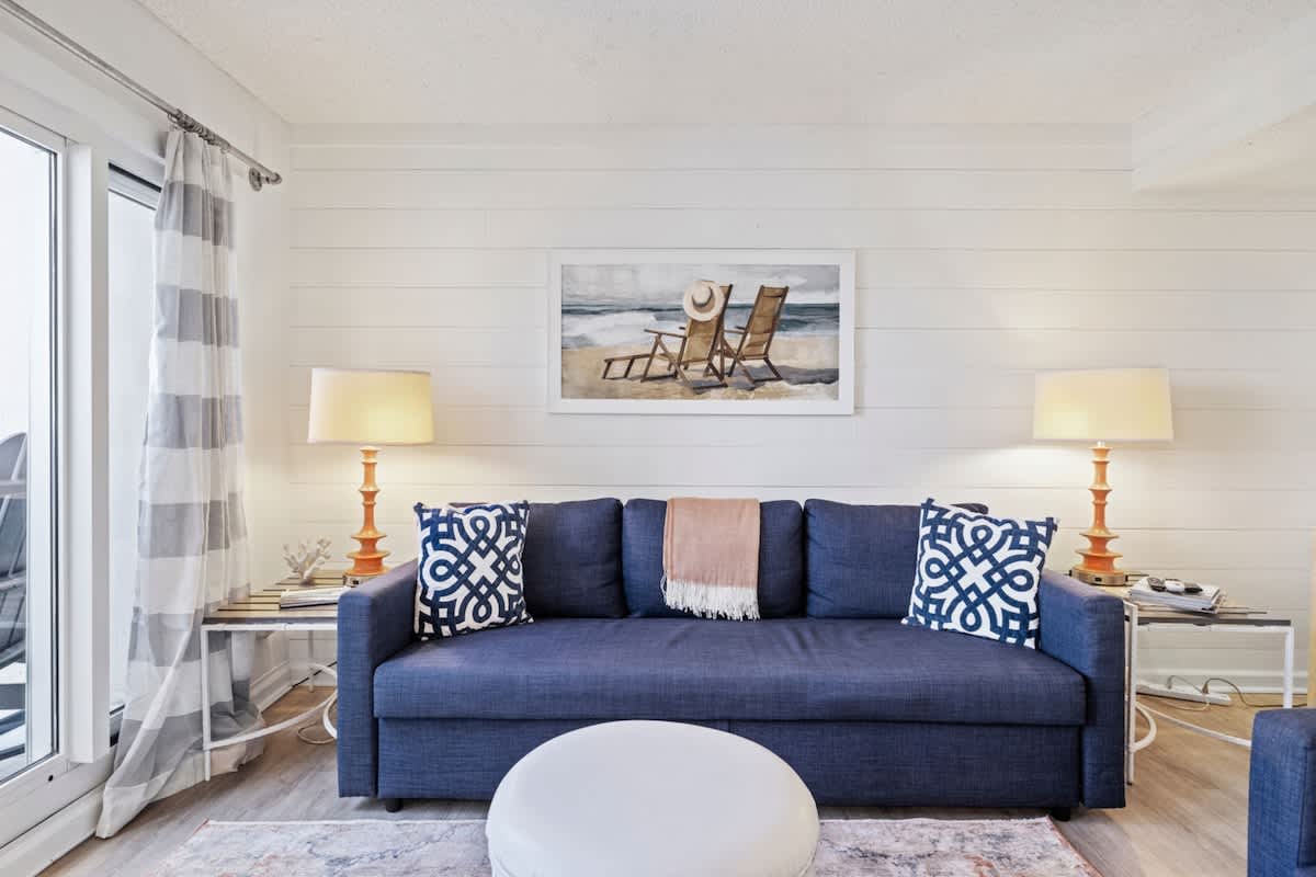 Pinnacle Port 304A – Cozy living room with blue couch, striped curtains, lamps, and beach-themed artwork.