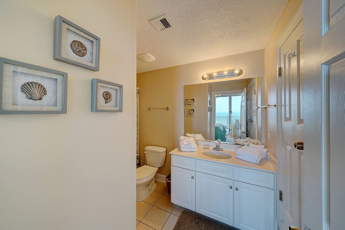 Ocean Villa 1104 – Bathroom with white cabinets, sink, toilet, and framed shell artwork; door leads to a balcony with ocean view.