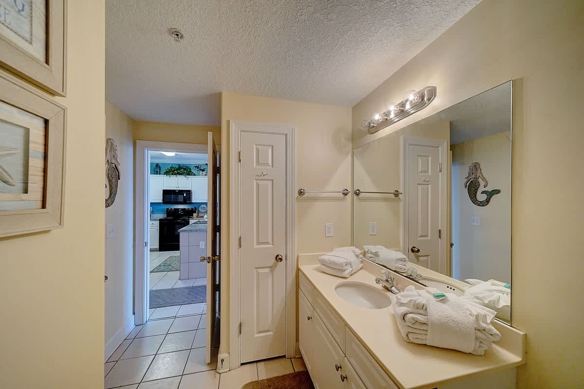 Ocean Villa 1104 – Bathroom with a sink, mirror, and towels; doorway leading to kitchen visible.