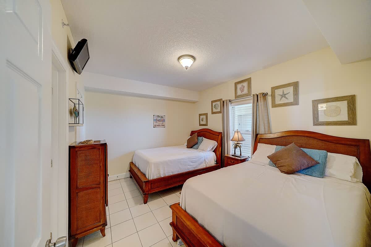 Ocean Villa 1104 – Guestroom with two beds, nightstand, lamp, dresser, and framed coastal decor on the walls.