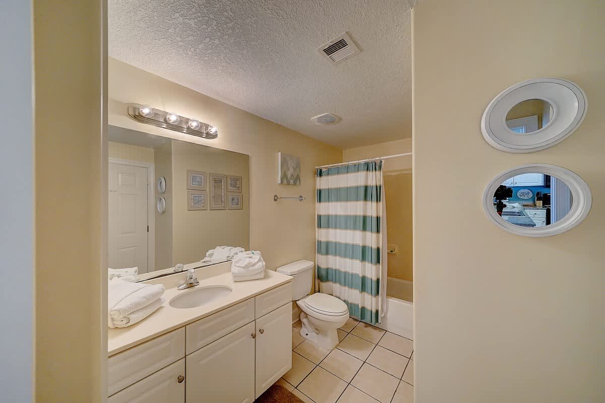 Ocean Villa 1104 – Bathroom featuring a sink, toilet, striped shower curtain, and framed art on a beige wall.