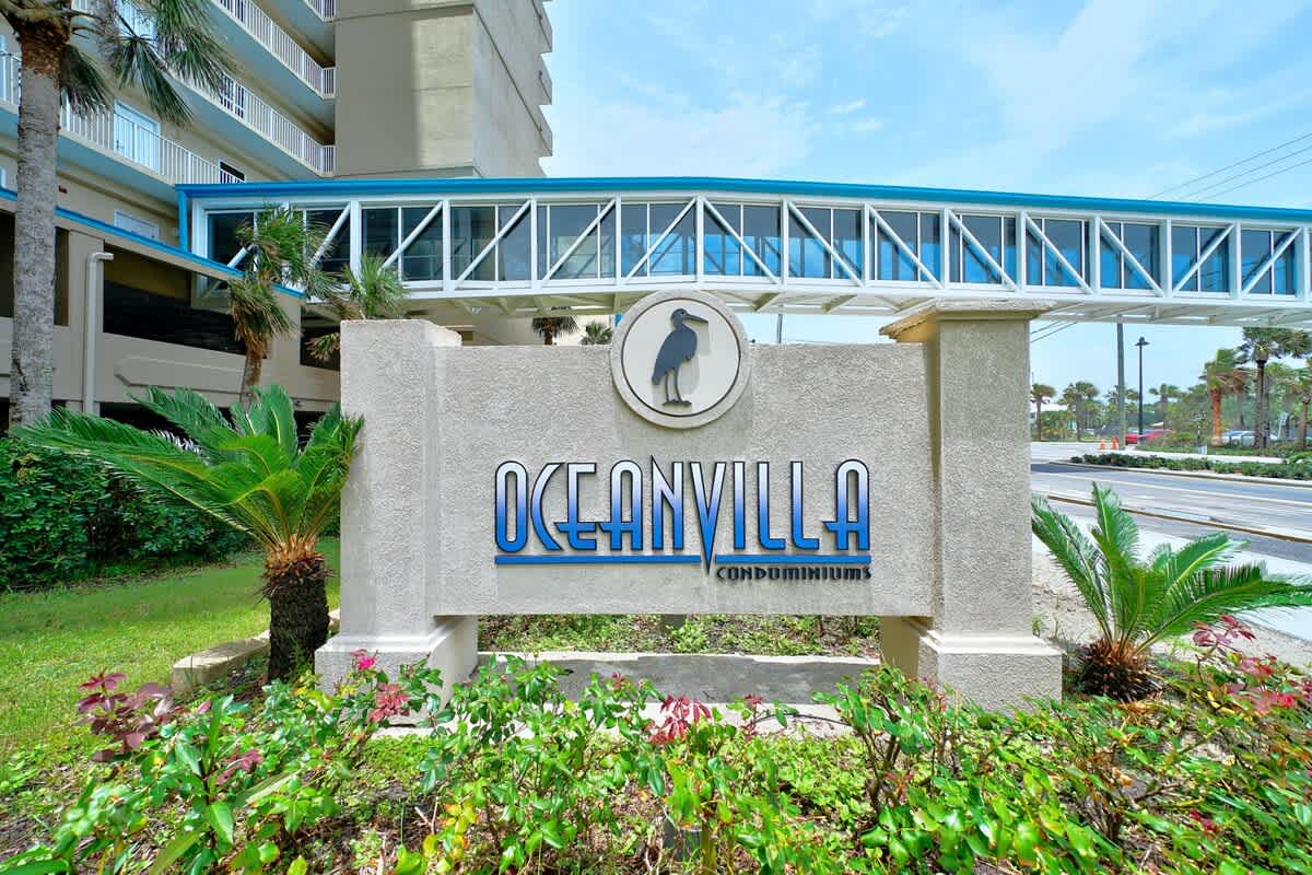 Ocean Villa 1104 – Sign for Ocean Villa condominiums with palm trees and a pedestrian bridge in the background.