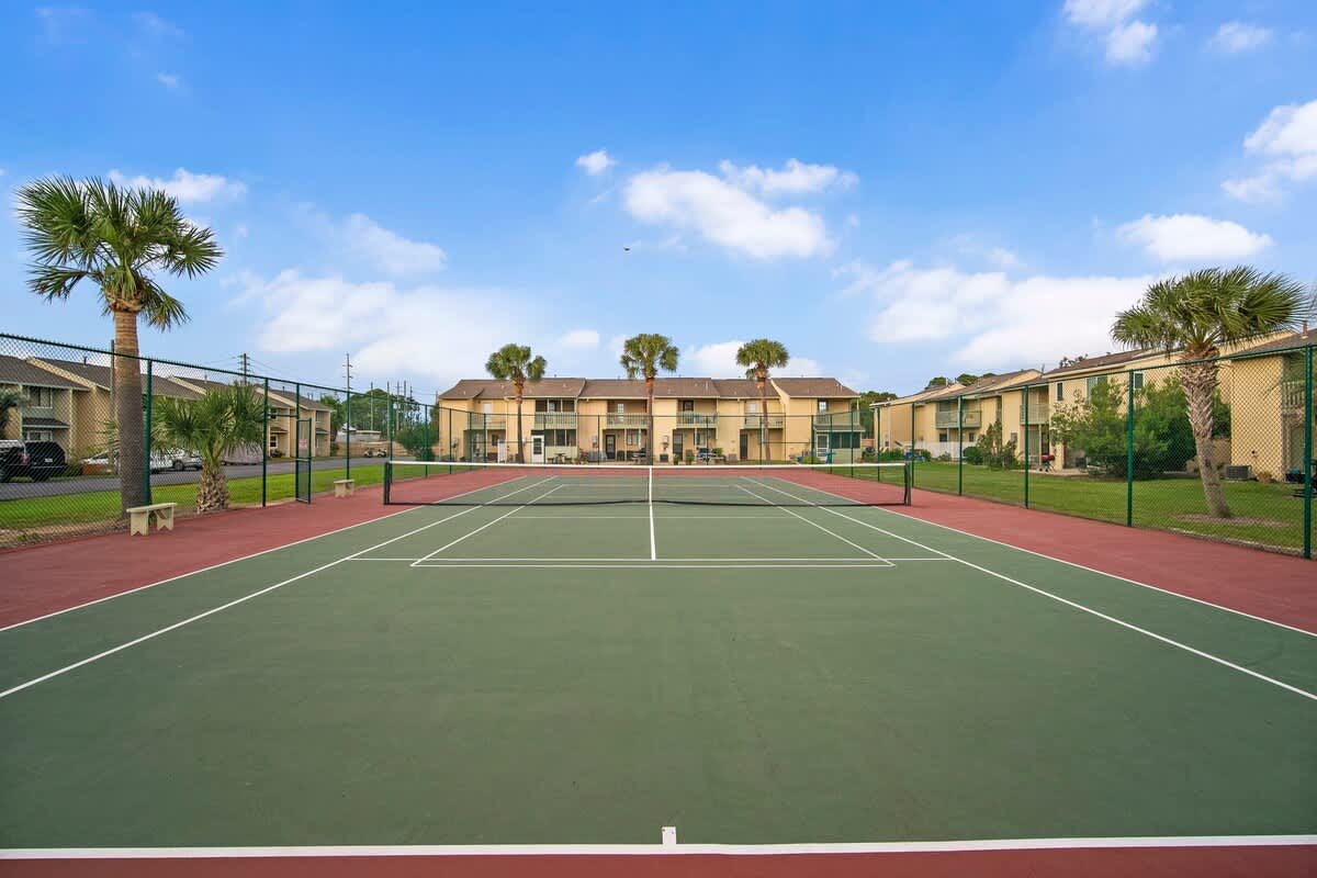 135 Grande Island Blvd – Tennis court with red and green surfaces, surrounded by palm trees and two-story buildings.