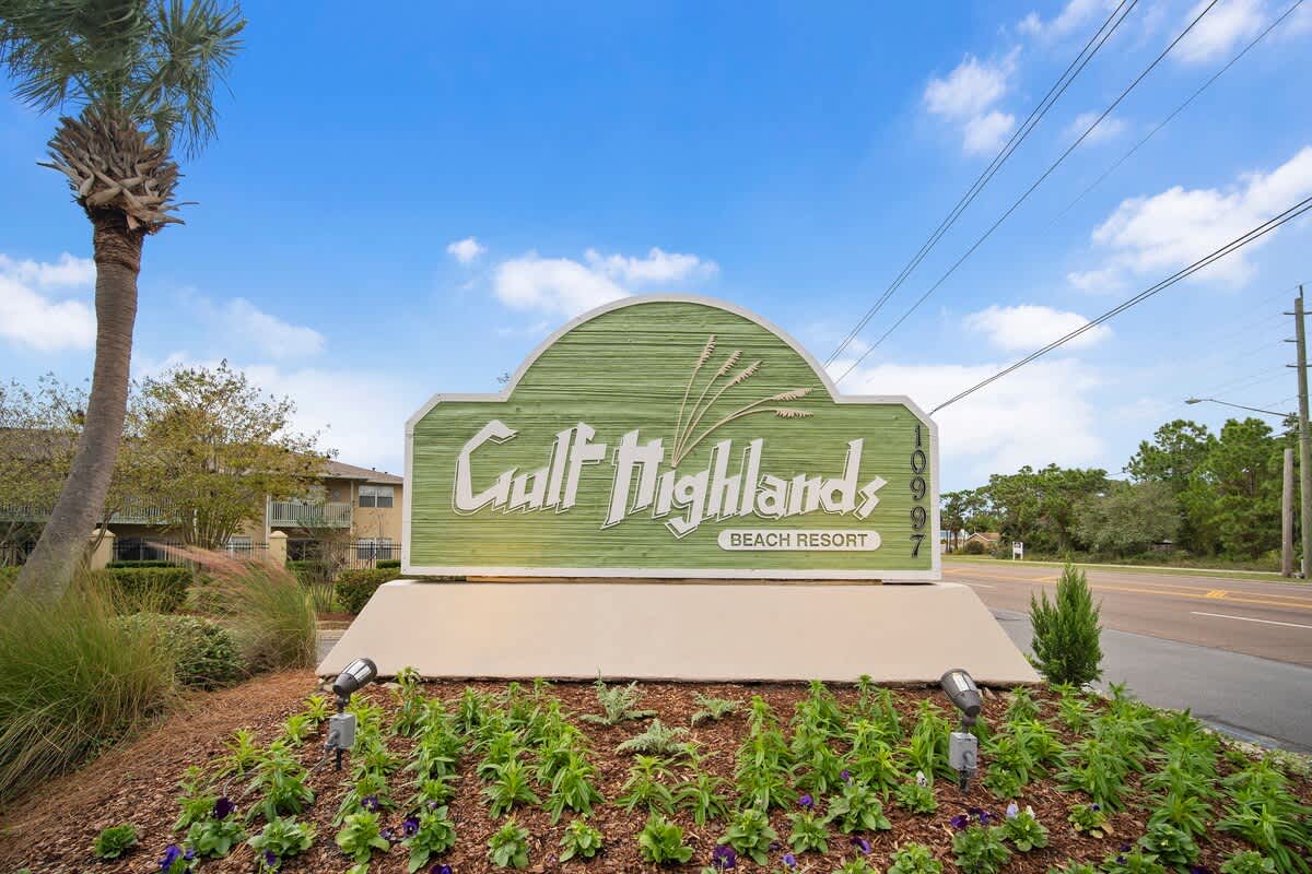 135 Grande Island Blvd – Gulf Highlands Beach Resort sign with landscaped garden, palm tree, and clear blue sky in the background.
