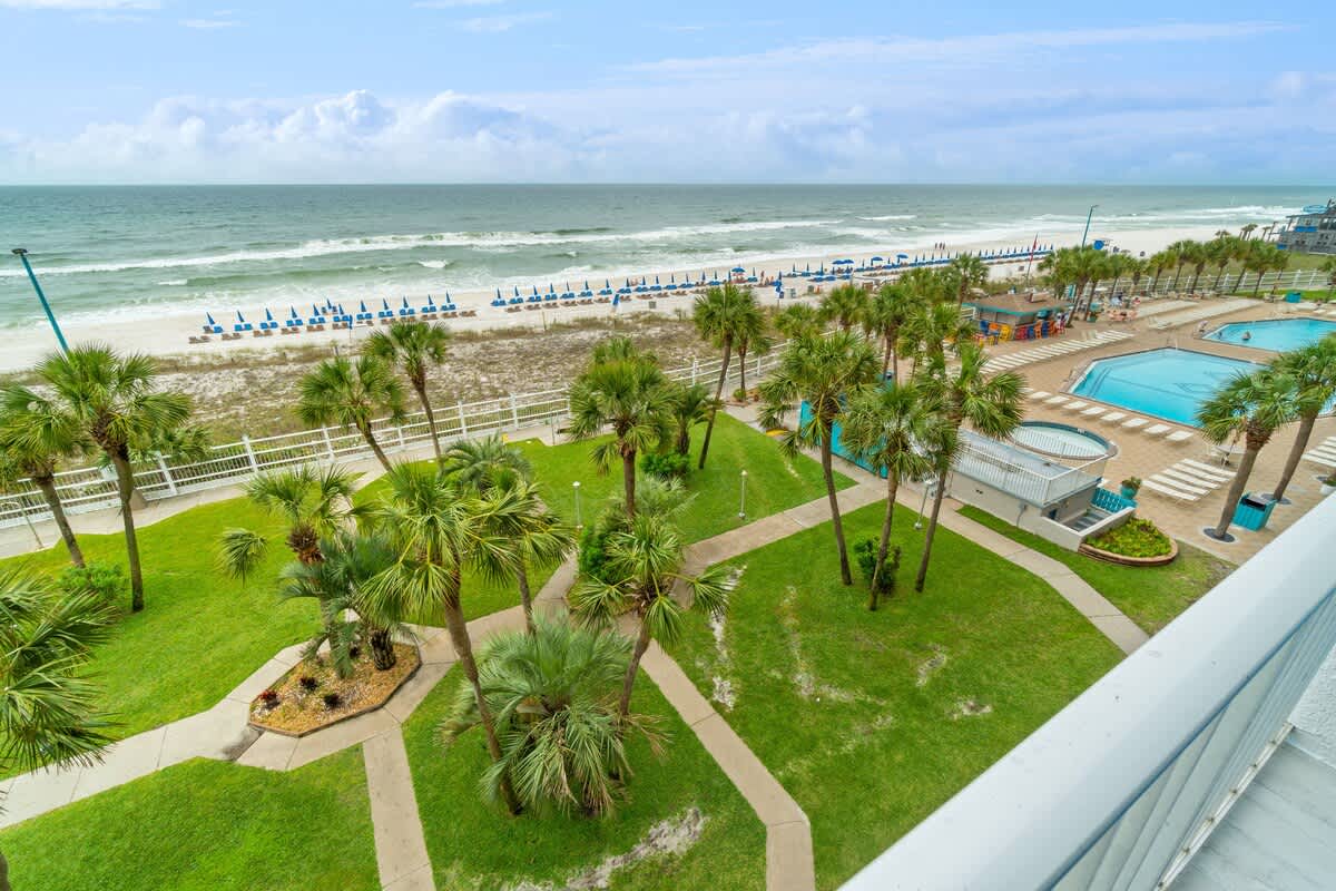 The Summit 529 – View from balcony showing green lawns, palm trees, pools, and beach chairs along the ocean.