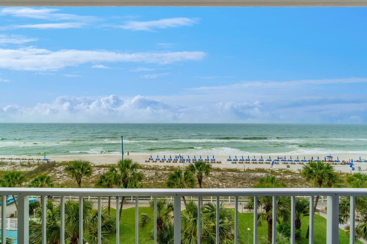 The Summit 529 – View from balcony overlooking beach, ocean, and lounge chairs under umbrellas. Palm trees in foreground.