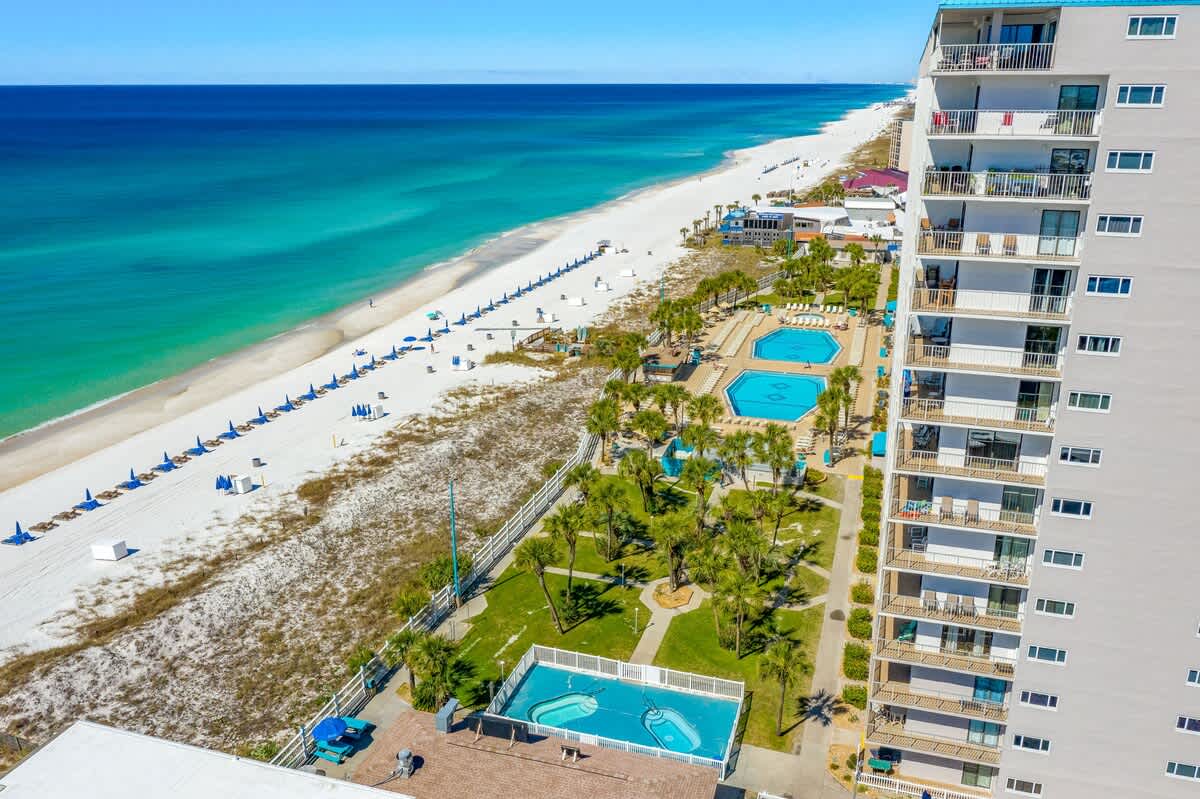 The Summit 927 – Aerial view of beachfront with pools, lounge chairs, and palm trees beside a high-rise building.