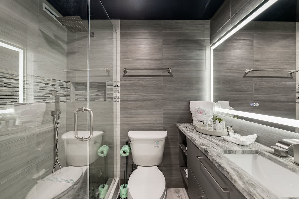 The Summit 1001 – Modern bathroom with gray tiles, glass shower, toilet, and large mirror with LED lighting.