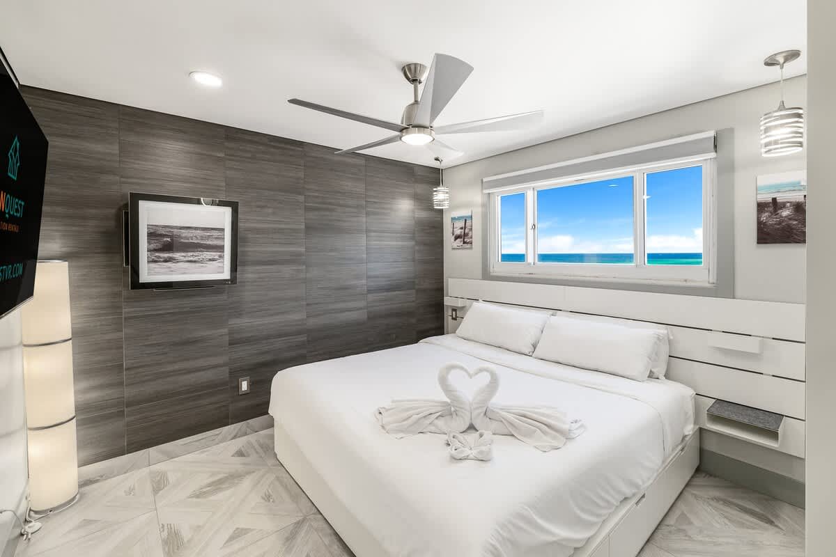 The Summit 1001 – Modern bedroom with a king bed, ceiling fan, TV, and ocean view through two windows.