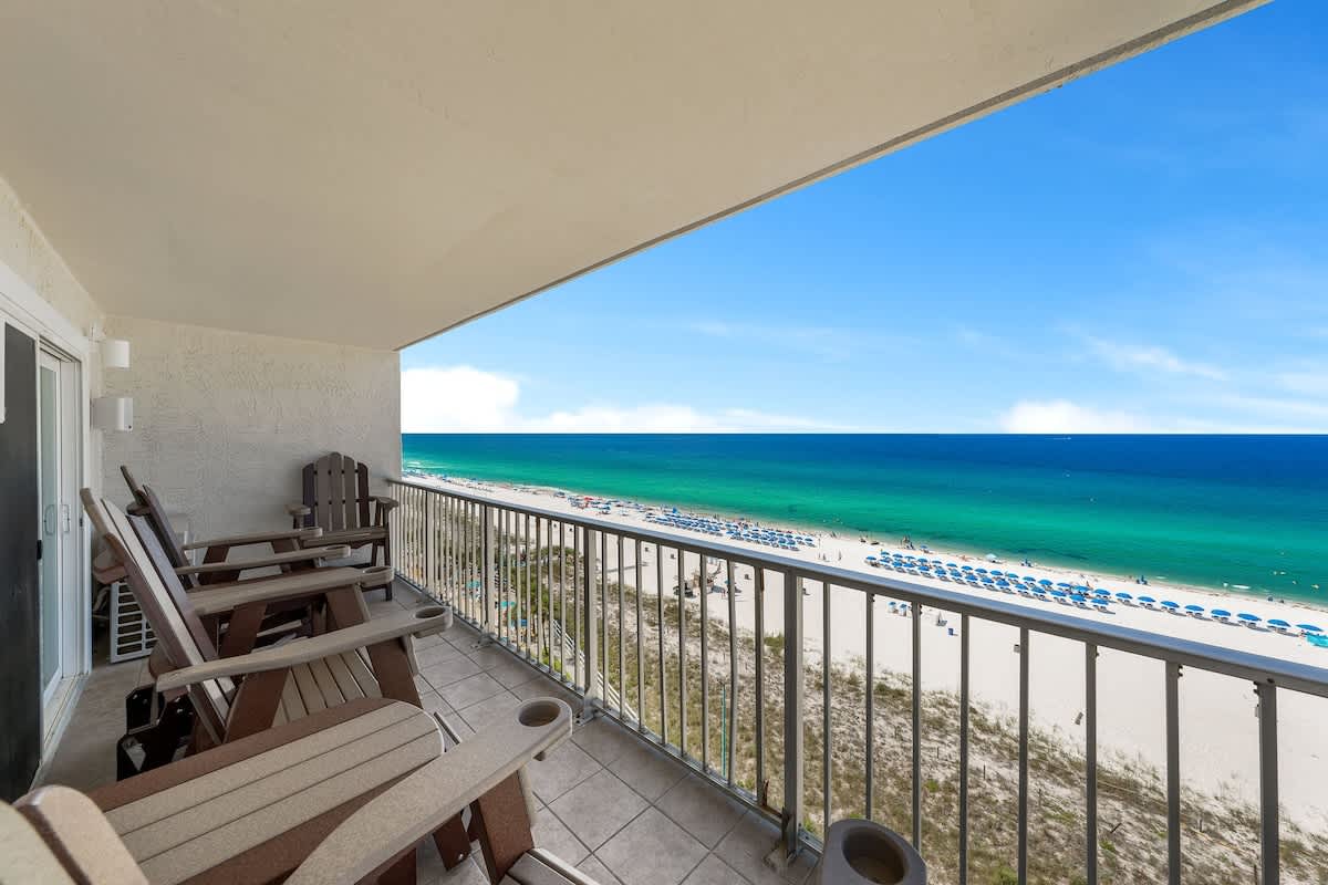 The Summit 1001 – Balcony view with wooden chairs overlooking a beach, turquoise water, and sun loungers on the sand.