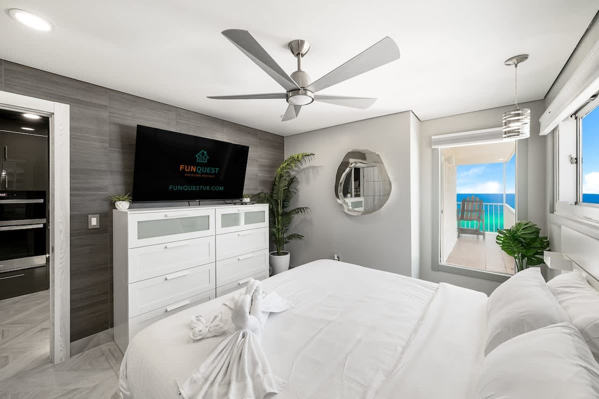 The Summit 1001 – Bedroom with ocean view, large window, white bedding, modern TV, and a ceiling fan.