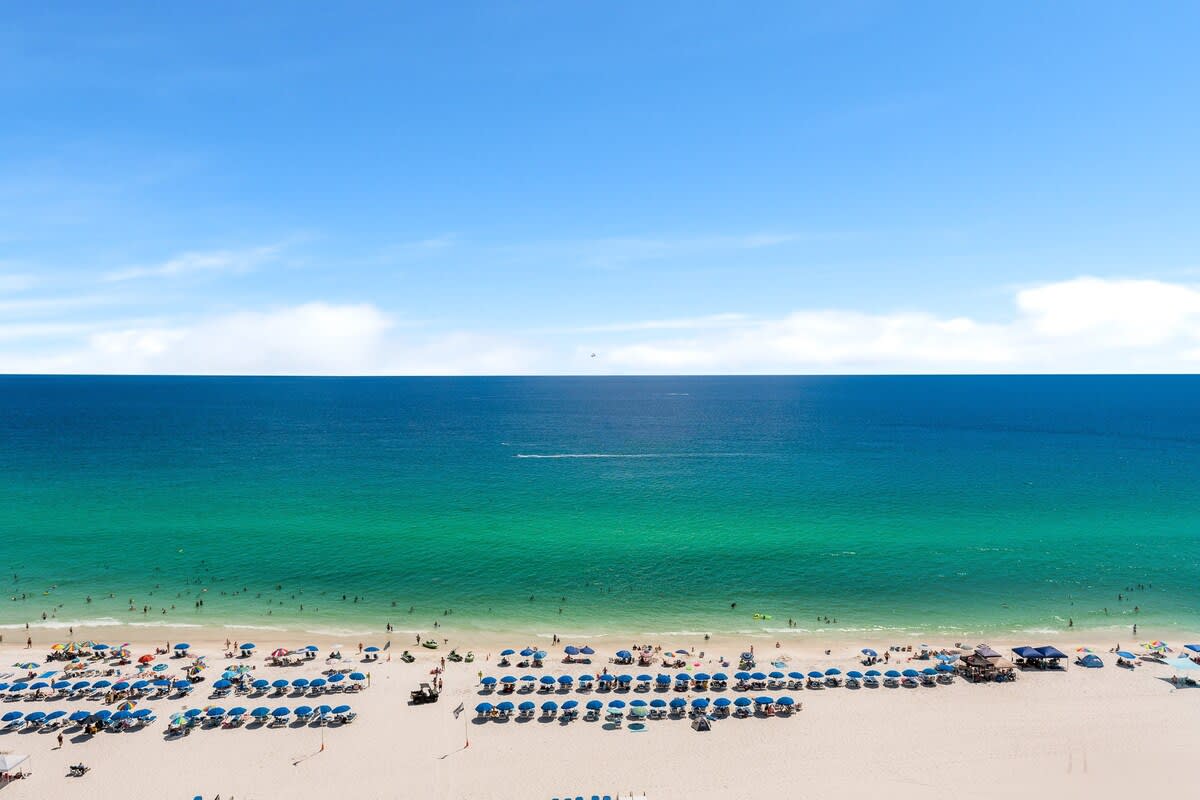 The Summit 1502 – Ocean view of a sandy beach with rows of umbrellas and people in the water.