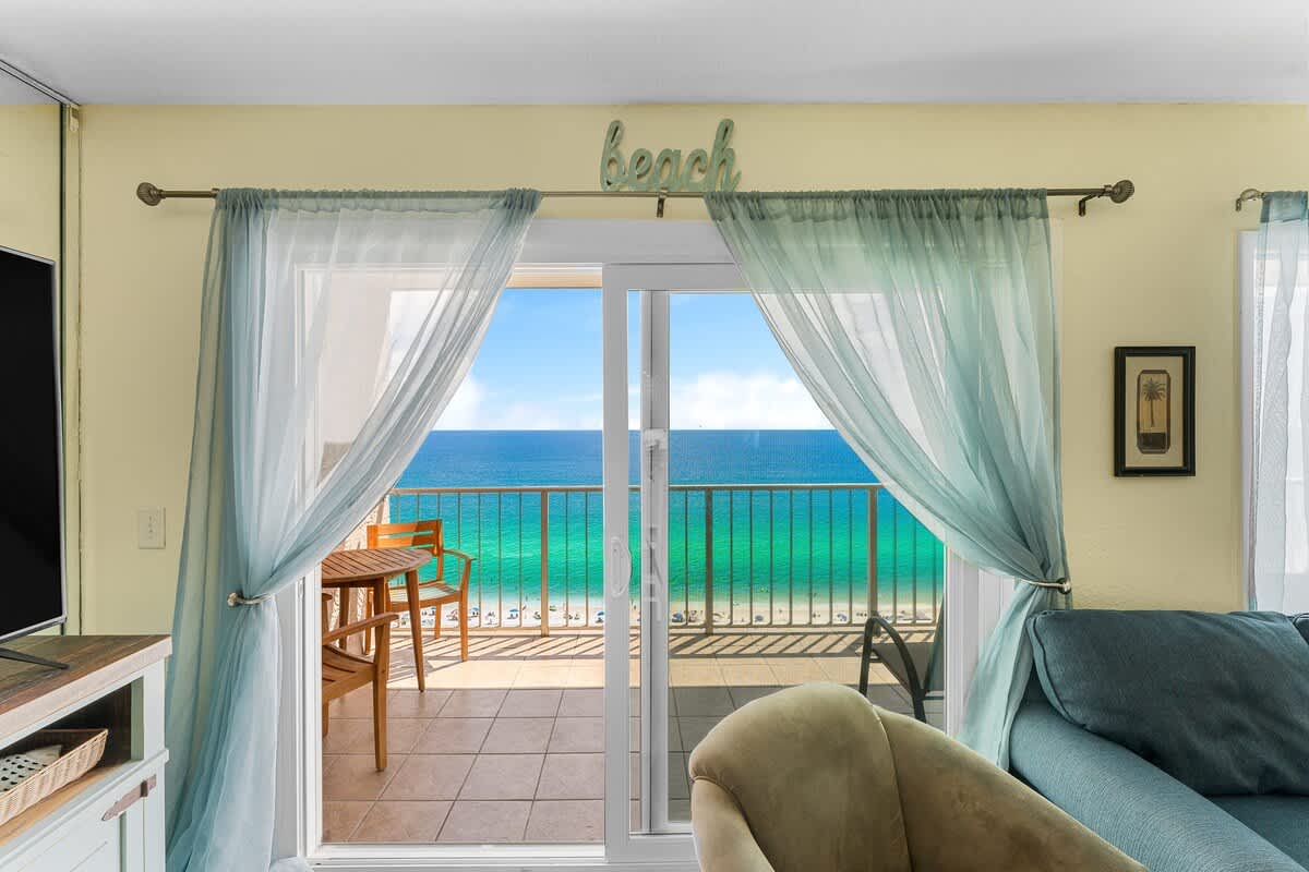 The Summit 1502 – Living room with balcony view of the ocean and beach, light curtains, and modern furnishings.
