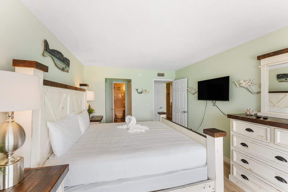 The Summit 1502 – Bedroom with a king bed, dresser, TV, and bathroom doorway in light green decor.