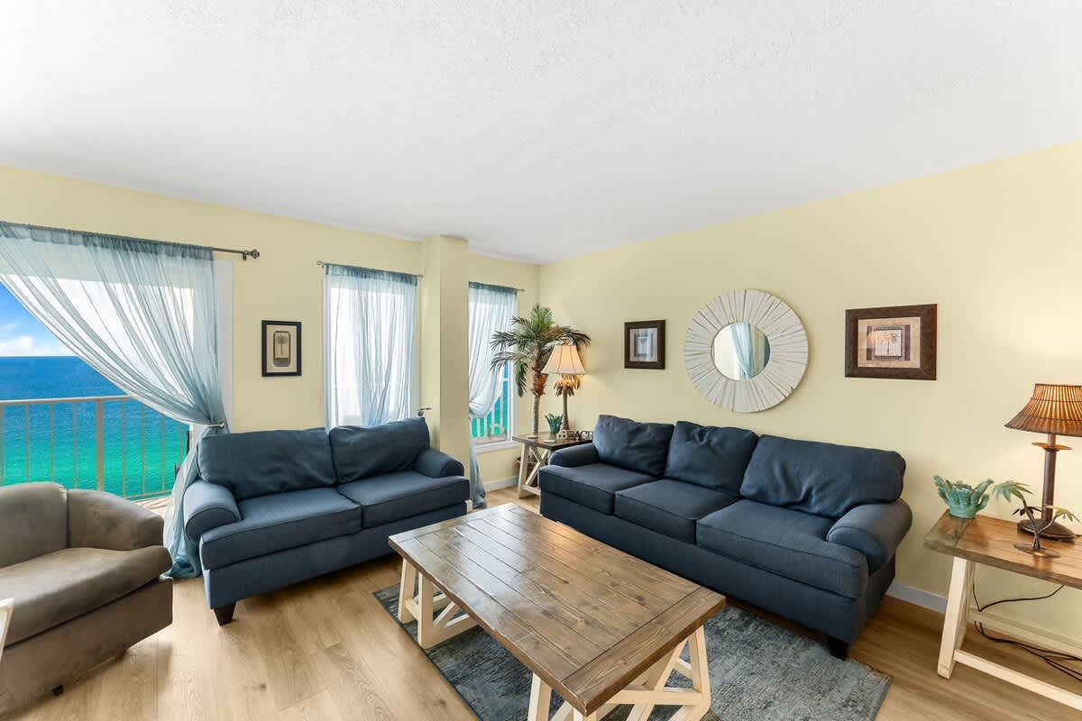 The Summit 1502 – Living room with two sofas, a coffee table, palm decor, and ocean view through sheer curtains.
