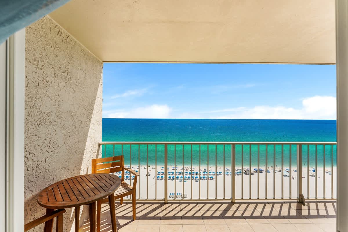 The Summit 1502 – Ocean view from a balcony with a wooden table and chairs, overlooking the beach and umbrellas.