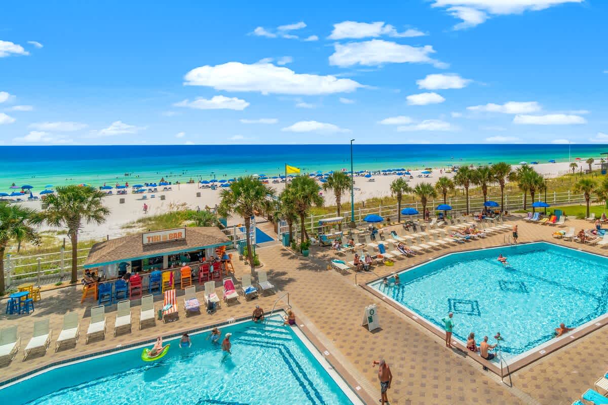 The Summit 828 – View of beachfront pools with lounge chairs, a tiki bar, and beach umbrellas under a clear blue sky.