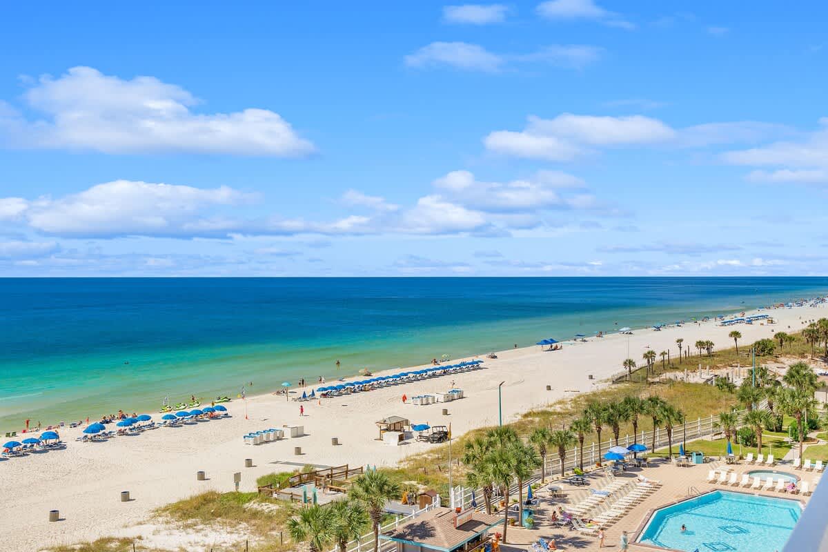 The Summit 828 – Elevated view of beach, pool area, and umbrellas along the shore under a blue sky.