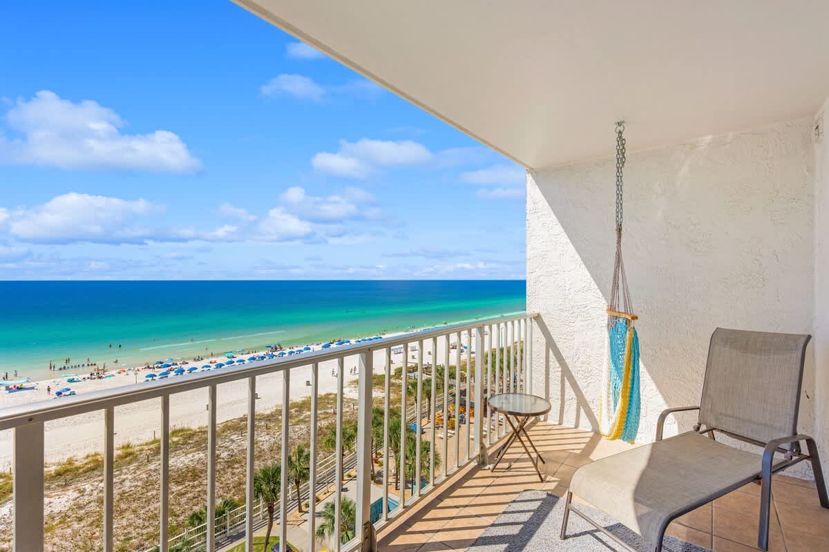 The Summit 828 – Balcony with a chair and hammock, overlooking a beach and ocean with umbrellas and people.
