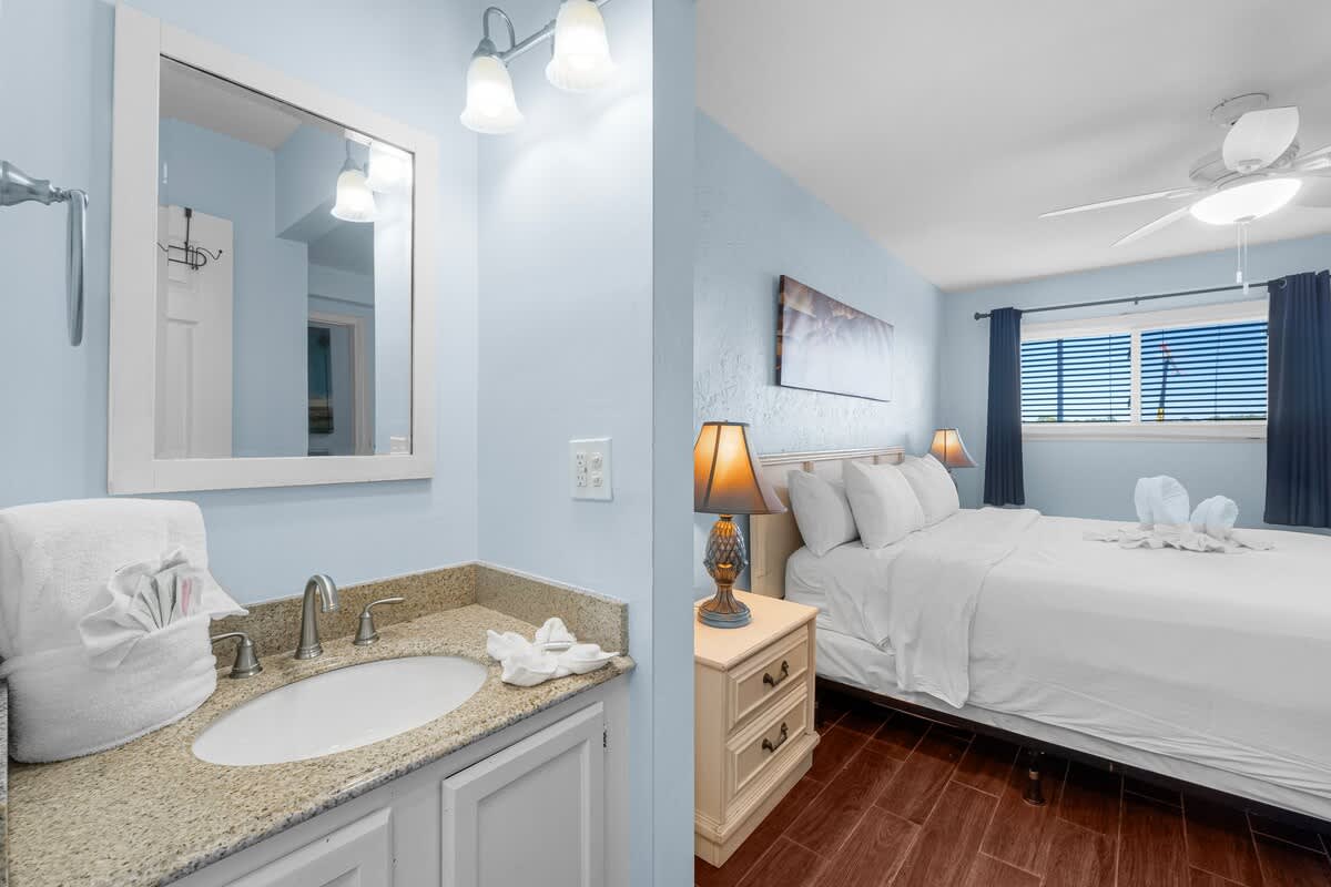 The Summit 408 – Bathroom with sink and mirror adjacent to a bedroom featuring a queen bed and window with curtains.