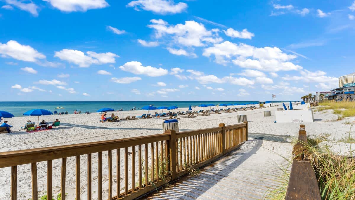 The Summit 408 – Boardwalk leading to a sandy beach with umbrellas and sun loungers under a blue sky.
