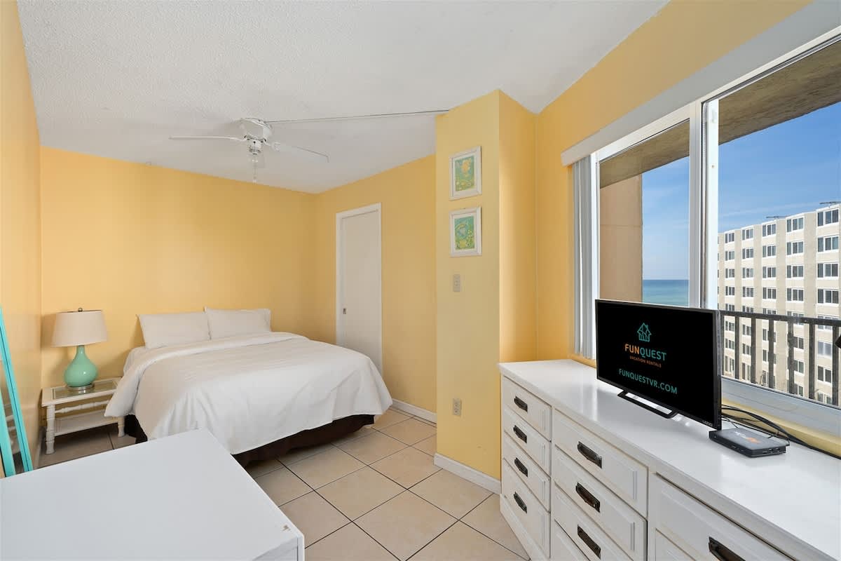 Moondrifter 606 – Bright bedroom with a queen bed, side lamp, and TV, featuring ocean views from large windows.