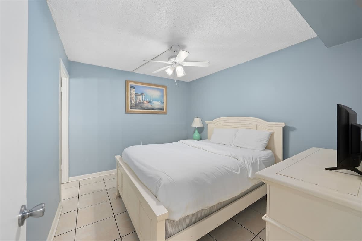 Moondrifter 606 – Bedroom with a white bed, blue walls, ceiling fan, lamp, and a small TV.