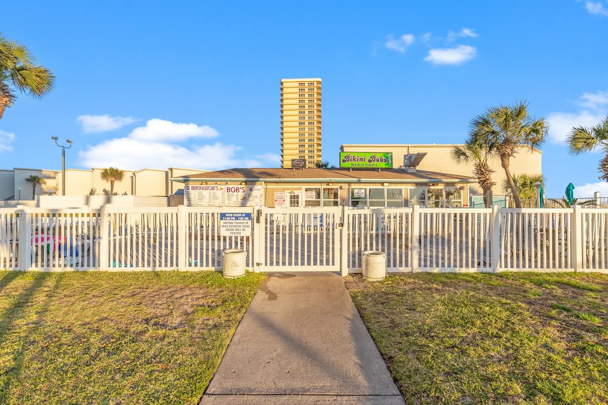 The Summit 524 – Bikini Bob's bar and grill with a fenced outdoor seating area and a tall building in the background.