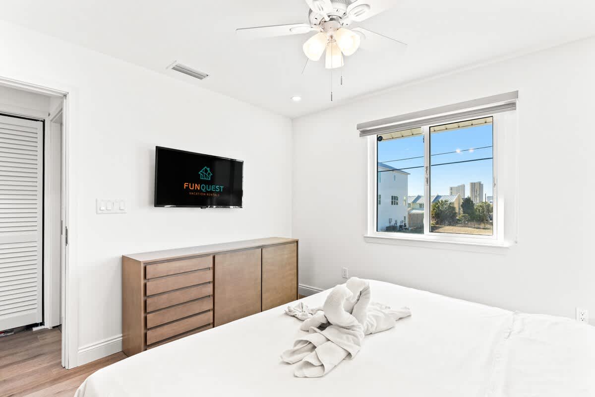 Ocean Breeze 2C – Bright bedroom with a queen bed, TV on the wall, and window view of buildings.