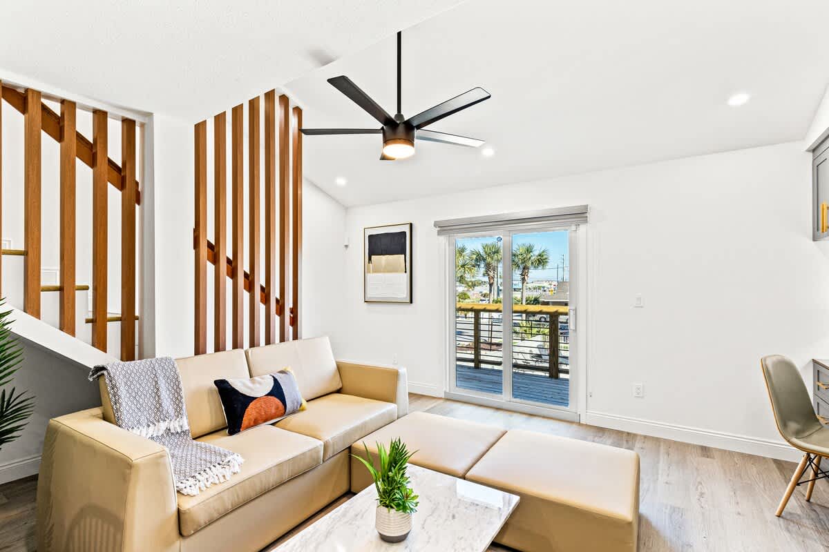 Ocean Breeze 2C – Living room with a beige sofa, sliding doors, wooden railing, and a view of palm trees outside.