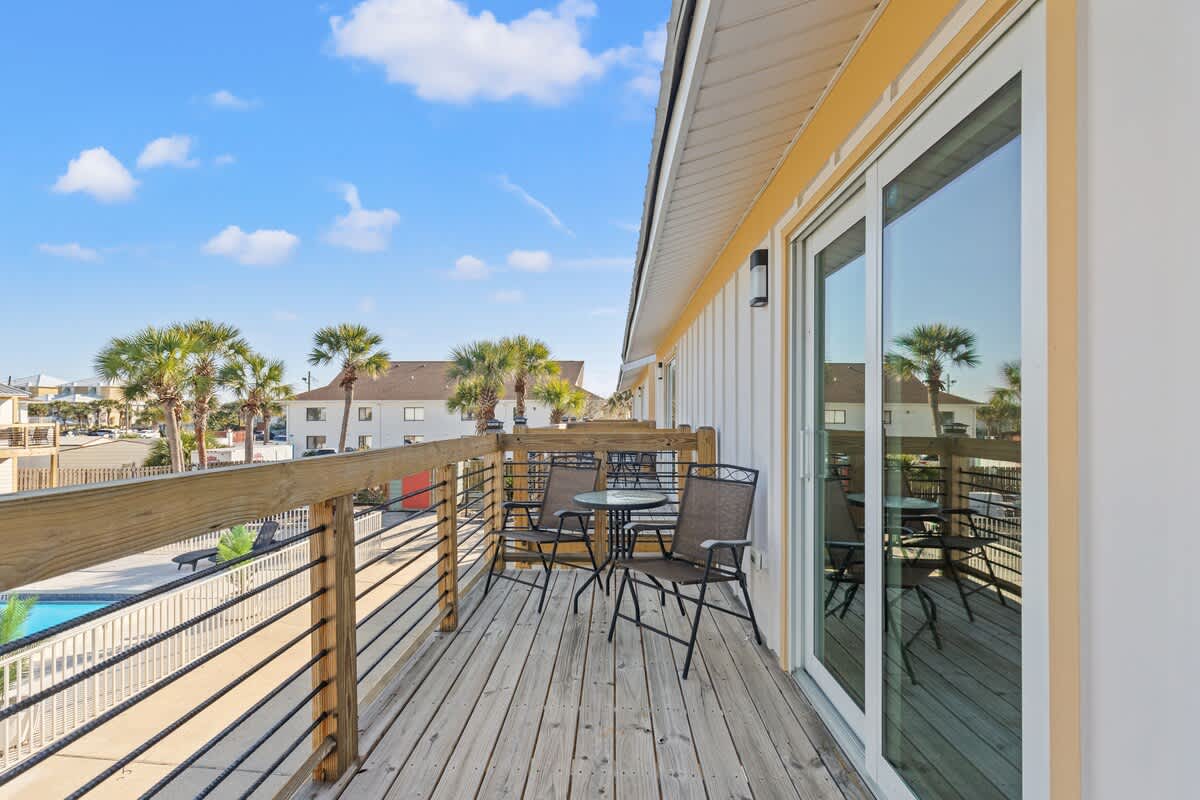 Ocean Breeze 2C – Balcony with wooden railing, two chairs, and a table overlooking palm trees and a pool.
