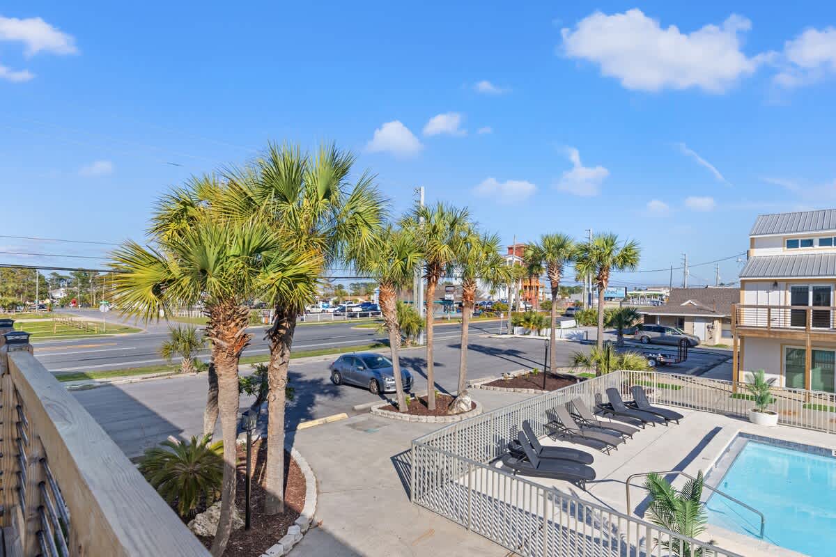 Ocean Breeze 2C – View from balcony with palm trees, pool area, and nearby parking lot. Blue sky in background.