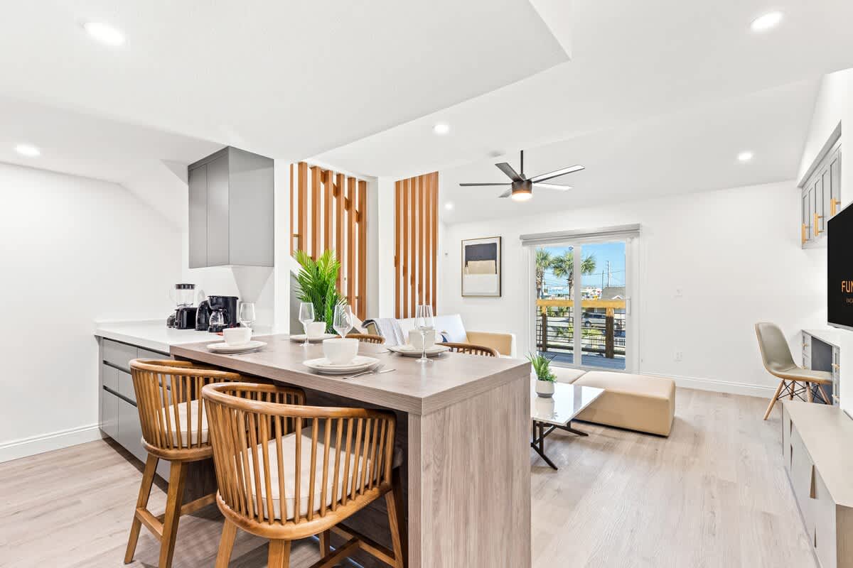 Ocean Breeze 2C – Modern living area with kitchen bar, wooden chairs, and a view of palm trees through a sliding door.