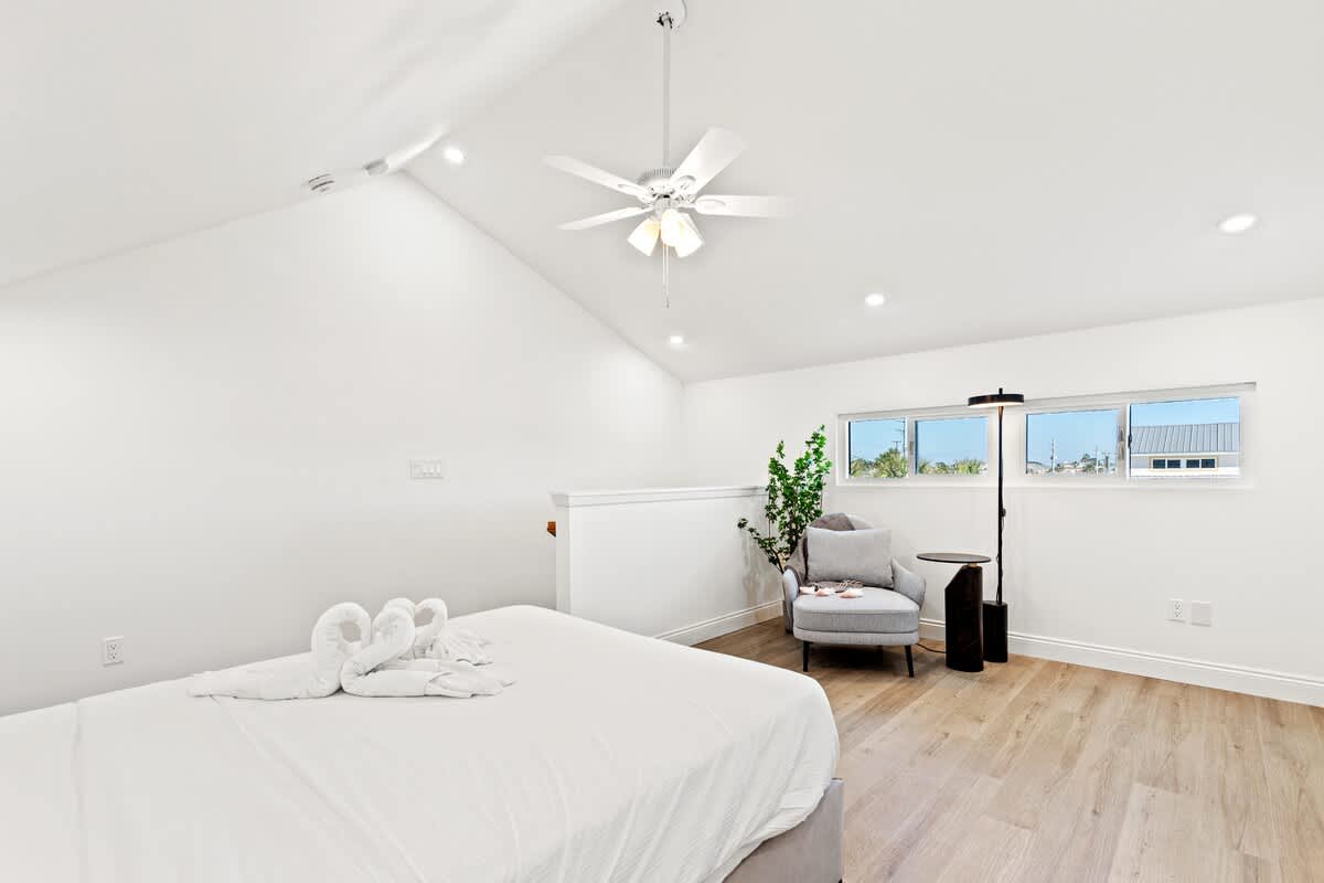 Ocean Breeze 2C – Bright bedroom with white walls, ceiling fan, queen bed, armchair, and windows showing outside view.