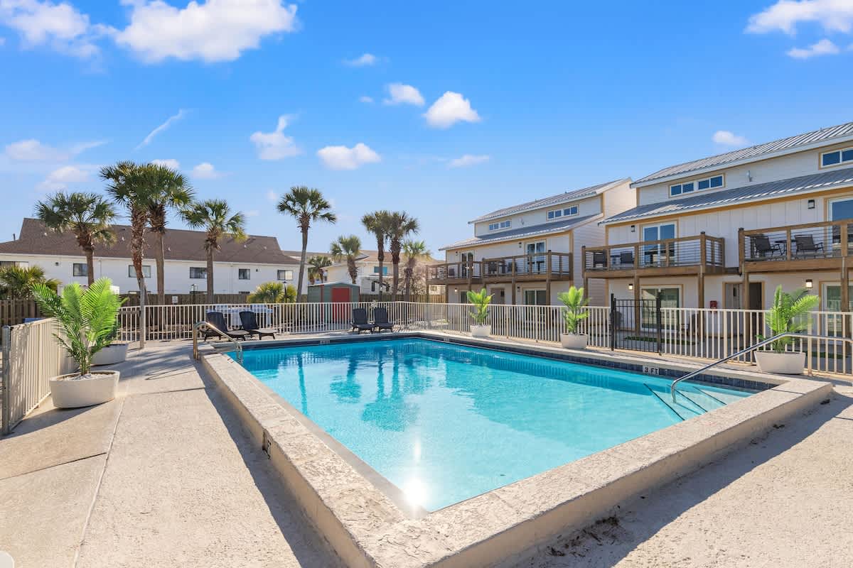 Ocean Breeze 3C – Pool area with lounge chairs, palm trees, and nearby rental buildings under a blue sky.