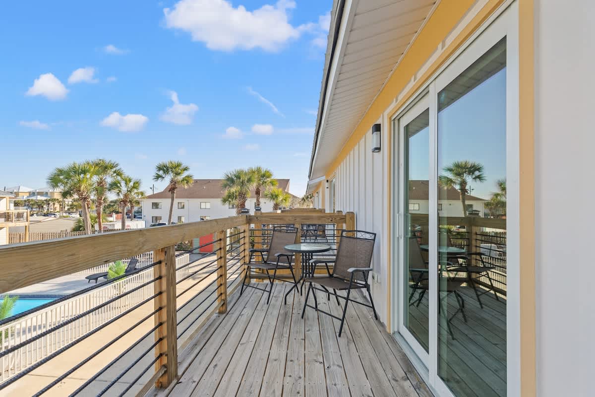 Ocean Breeze 3C – Wooden balcony with seating, overlooking a pool area and palm trees under a clear blue sky.