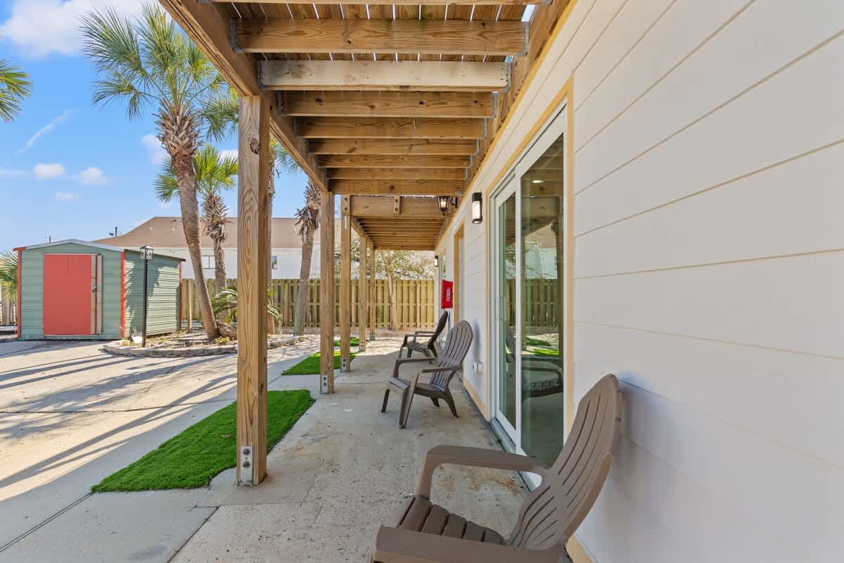 Ocean Breeze 5B – Covered patio with two chairs, small turf area, and view of palm trees and storage shed.