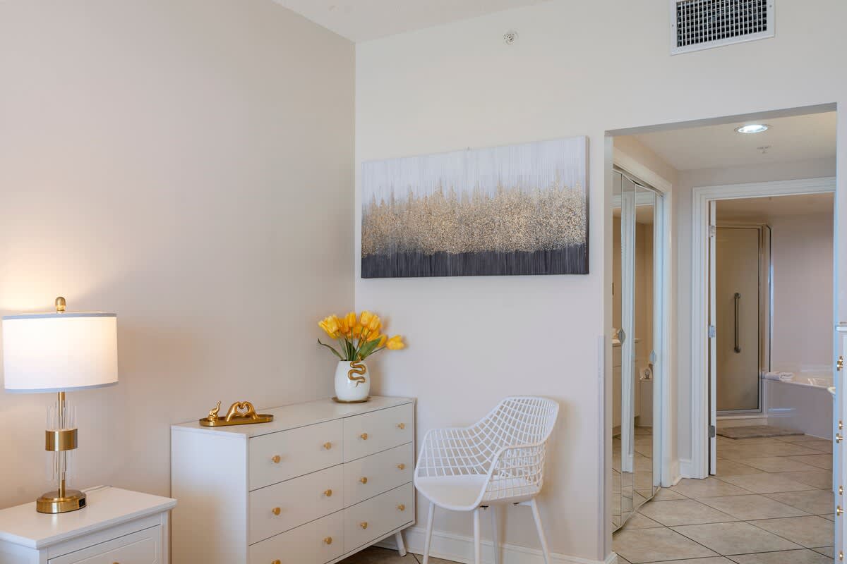 Majestic T2 - 1606 – Entryway with white dresser, chair, lamp, and artwork; bathroom door visible in the background.
