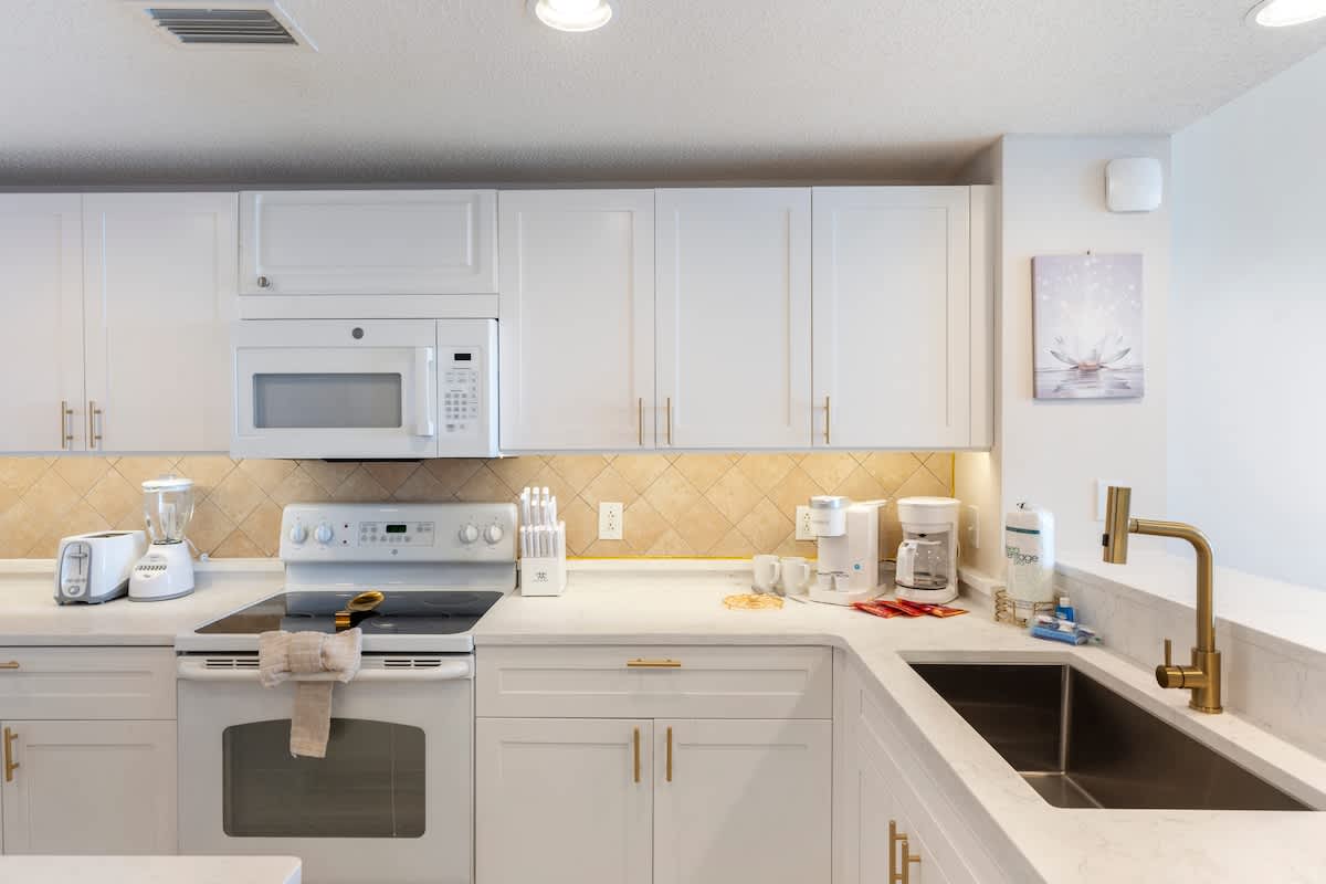 Majestic T2 - 1606 – Modern kitchen with white cabinets, appliances, and gold accents; includes sink, stovetop, and countertop.
