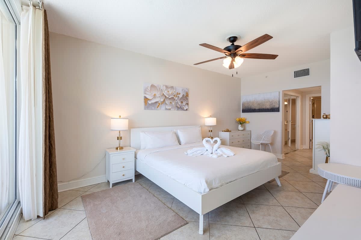 Majestic T2 - 1606 – Bright bedroom with a large bed, two lamps, and wall art, plus a view into a hallway.