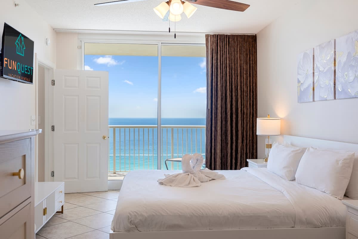Majestic T2 - 1606 – Bedroom with white bed, balcony view of the ocean, and decorative towel art.