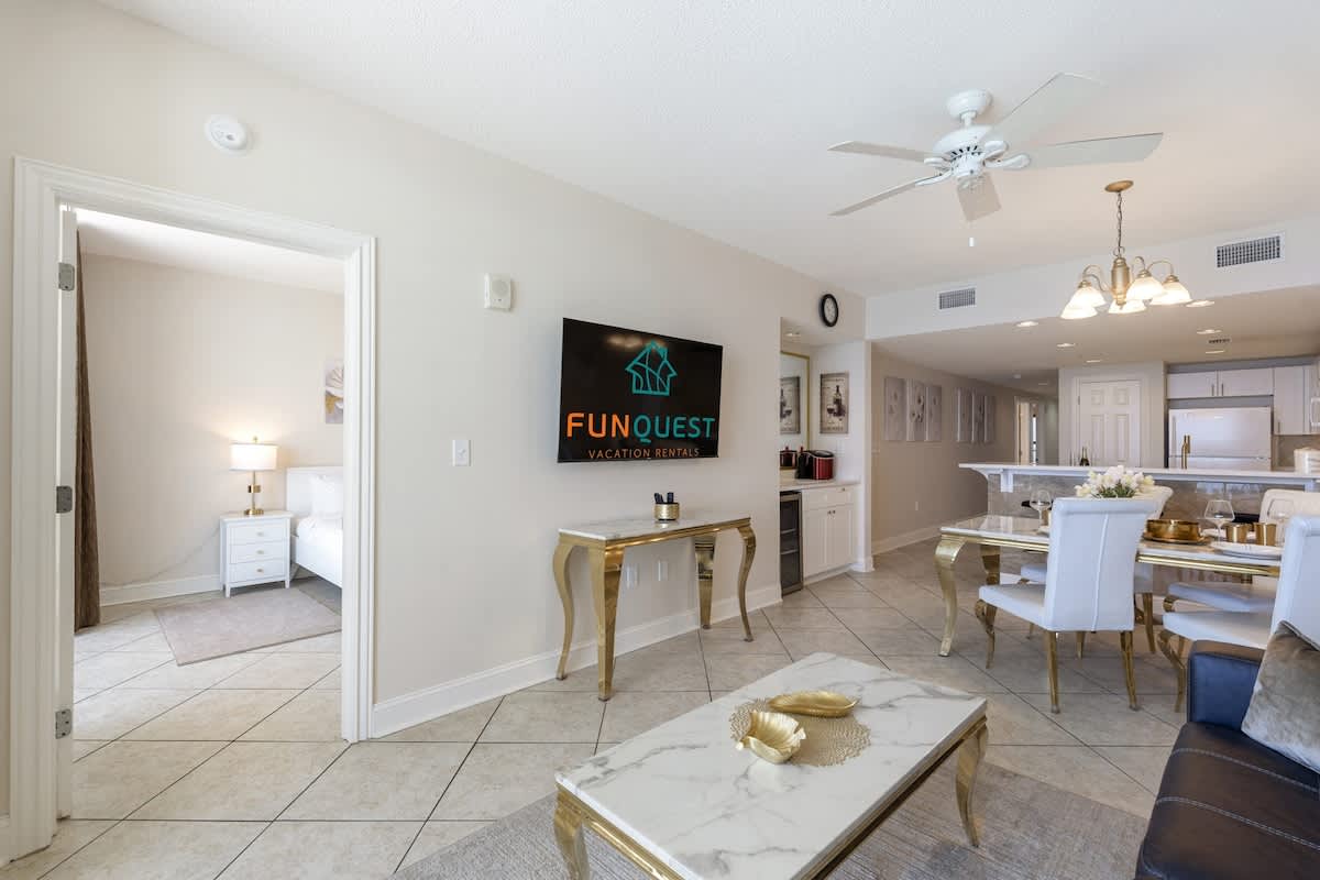 Majestic T2 - 1606 – Living room with TV, dining area, and view into adjacent bedroom with lamp and white bed.