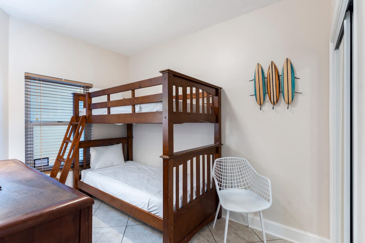Majestic T2 - 1606 – Bunk bed with a white chair, window with blinds, and surfboard wall decor in a small room.