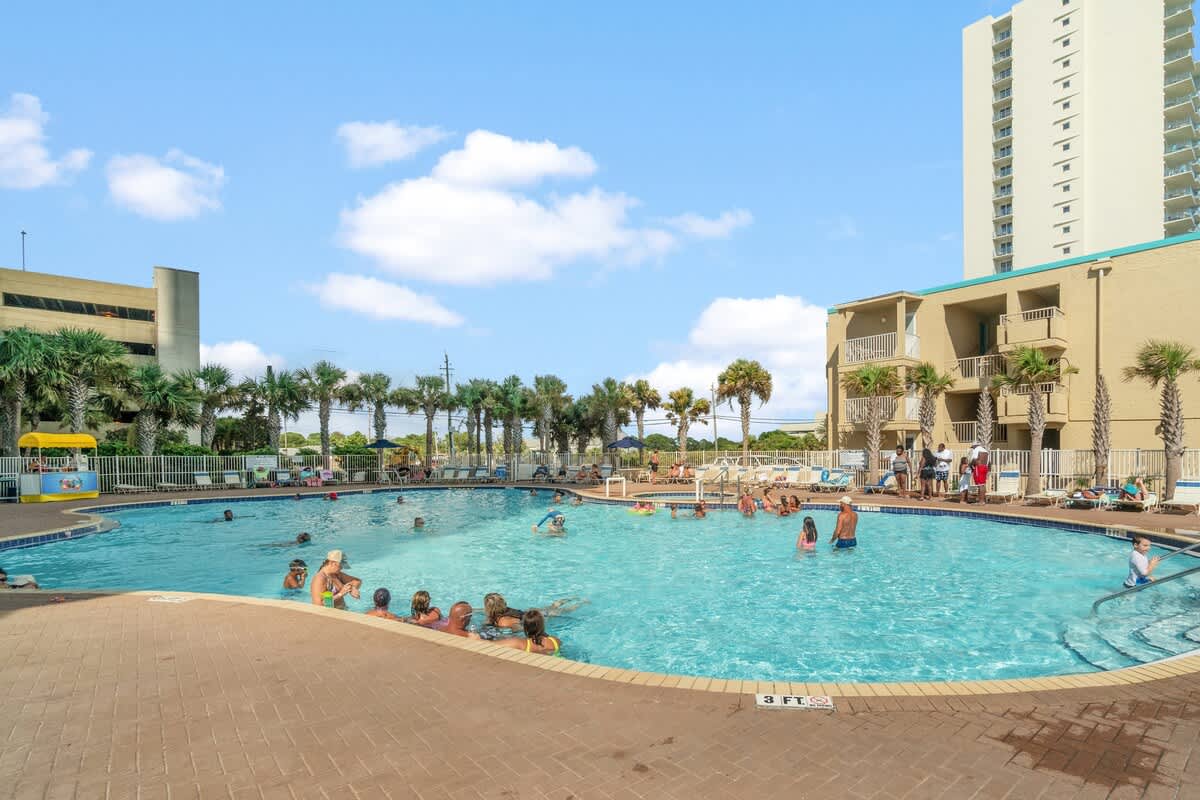 Tidewater 2615 – Swimming pool area with people, palm trees, and adjacent building in a vacation rental setting.