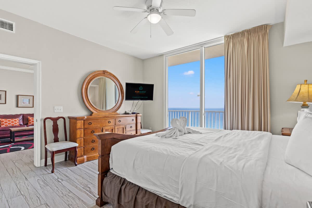 Tidewater 2615 – Bright bedroom with ocean view, dresser, chair, TV, and bed with towel art.