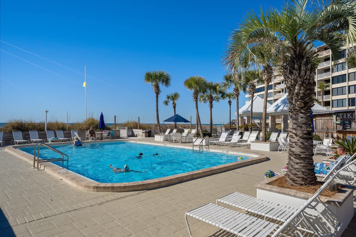 Dunes C605 – Pool area with chaise lounges, palm trees, and ocean view under a clear blue sky.