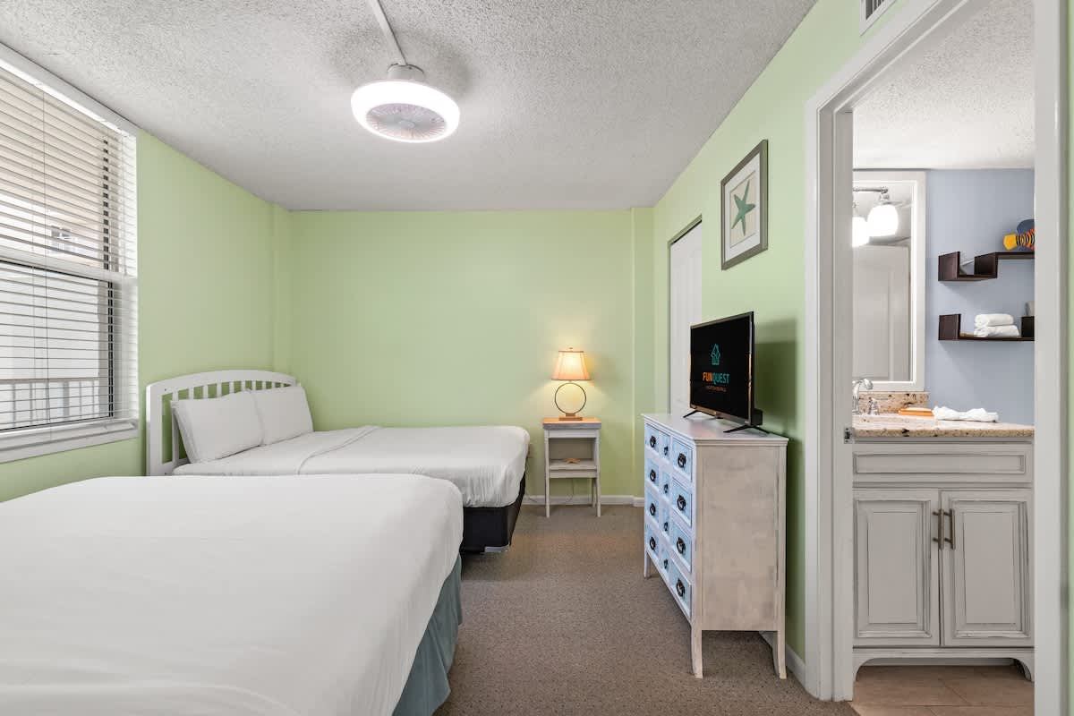 Dunes C605 – Bedroom with two beds, television, dresser, lamp, and adjoining bathroom in green-painted room.