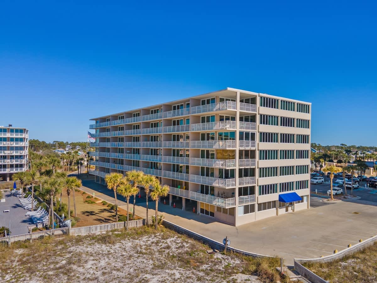 Dunes C605 – Five-story beachfront building with balconies, surrounded by palm trees and parking area. Clear blue sky.
